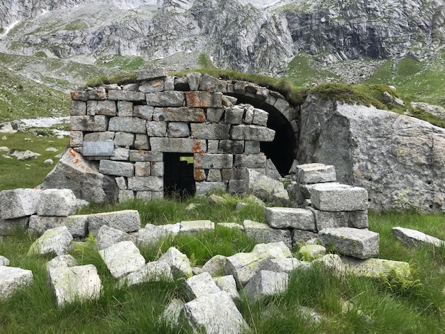 Foto di un piccolo rifugio costruito da rocce di fronte ad una montagna e circondato da piante verdi