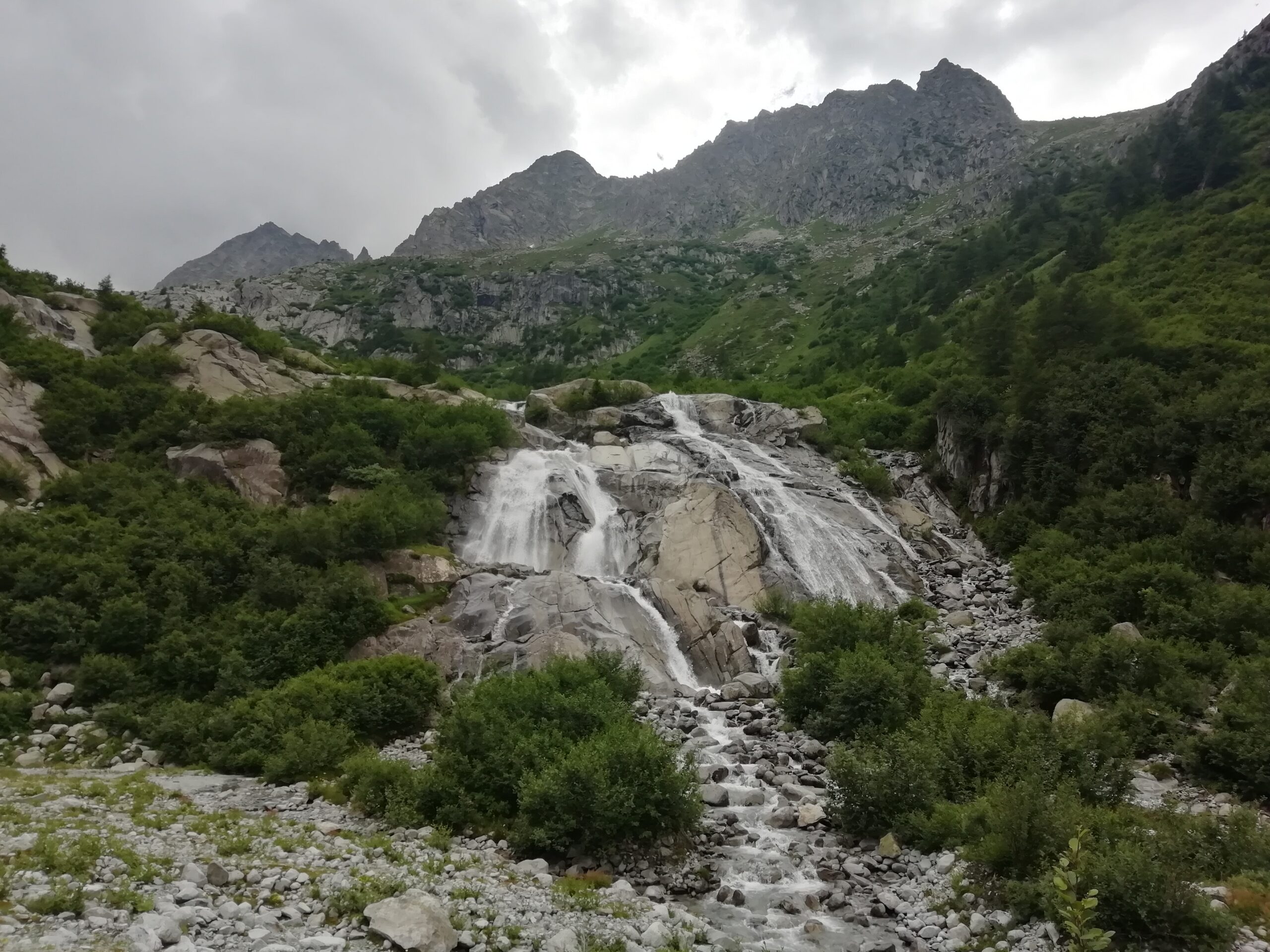 Foto di una piccola cascata ornata da piccole e grandi rocce, da piante verdi e da montagne macchiate dal verde